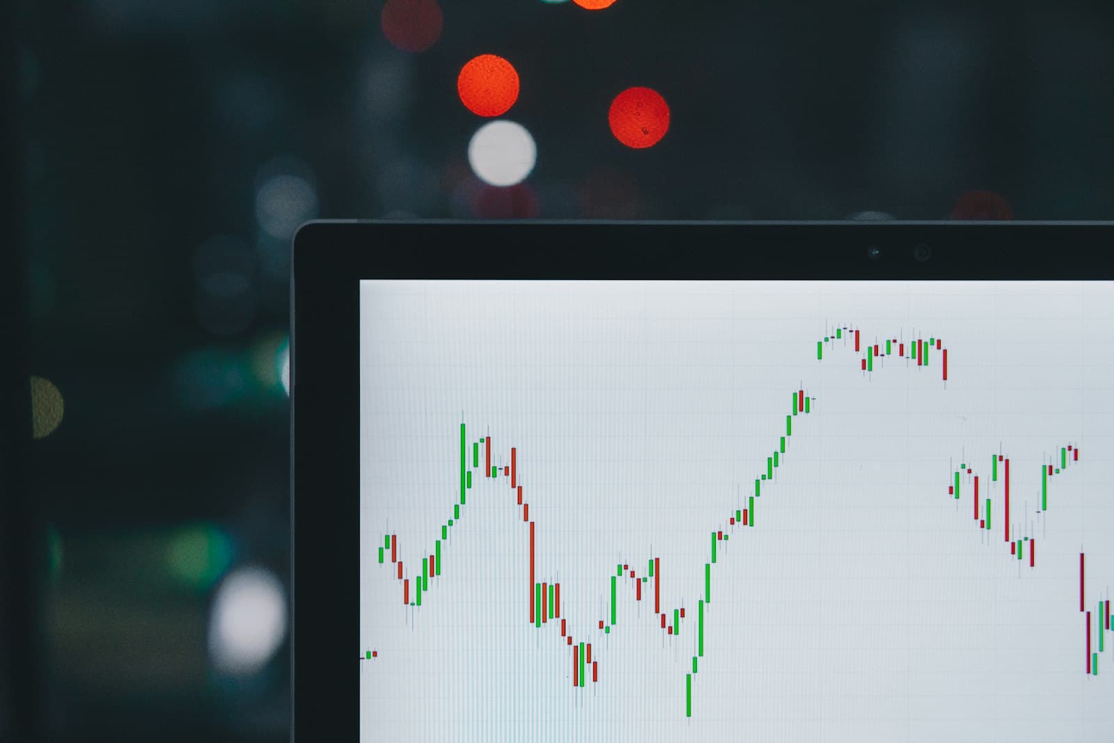 A laptop screen showing a candlestick price chart with green and red candles, with blurred city lights in the background