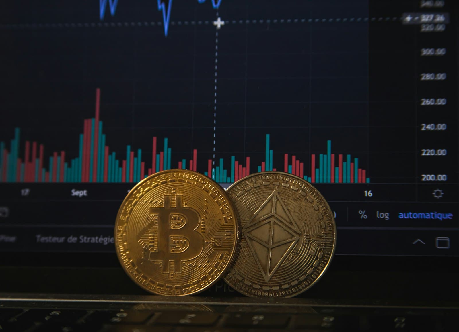 A gold bitcoin coin and a silver ethereum coin standing upright in front of a screen showing a price chart with volume bars